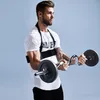 A man is performing bicep curls with a barbell while wearing a black arm blaster. Produktbild-Vorschau