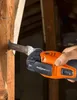 A person's hand is holding an orange and black oscillating multi-tool, using it to cut into a wooden beam in what appears to be an attic or unfinished room. Náhľad obrázku produktu