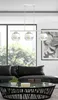 A modern living room with a black leather sofa, a glass coffee table with decorative items, and a large window with a view of trees. A white pendant light fixture hangs above the coffee table. Podgląd zdjęcia produktu