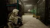 A person in a white suit crouches behind a blue industrial cart in a dimly lit warehouse aisle. Náhľad obrázku produktu
