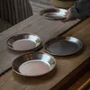 Several metal plates are arranged on a wooden table, with a hand holding one plate above the others. Náhled obrázku produktu