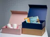 Two open gift boxes, one pink and one blue, are displayed on a surface against a light gray background. The pink box contains two white mugs and some pink filler material. The blue box contains two books and is placed on top of a brown box. Podgląd zdjęcia produktu