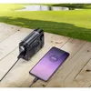A portable radio is charging a smartphone on a wooden surface, with a grassy field and water in the background. Produktbild-Vorschau