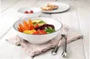 A white dessert bowl filled with colorful food, placed on a pink napkin on a wooden table. Another bowl and a plate with bread are in the background. Anteprima dell’immagine prodotto