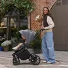 A woman is pushing a baby stroller on a sidewalk in front of a building with plants and a brick facade. Náhled obrázku produktu