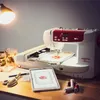 A sewing and embroidery machine is set up on a wooden desk with a tablet displaying a design, thread spools, scissors, and a measuring tape. Produktbild-Vorschau