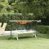 A rotisserie grill is set up in a backyard with lush green trees and hedges. Náhled obrázku produktu