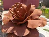 A close-up of a rusty metal flower sculpture with a pond and greenery in the background. Produktbild-Vorschau