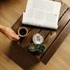 A hand holding a cup of coffee next to a newspaper, a candle, and small succulents on a wooden crate. Náhled obrázku produktu