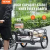 A person is riding a bicycle with a trailer attached, loaded with items, on a paved path surrounded by greenery. Podgląd zdjęcia produktu