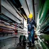 A man in a yellow hard hat is using a vacuum cleaner to clean up a wet floor in a room with many pipes. Náhled obrázku produktu