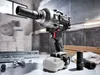 A cordless impact wrench with a socket attached, placed on a workbench with other tools and materials in the background. Produktbild-Vorschau