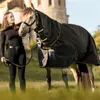 A woman stands next to a horse wearing a turnout rug, with a building in the background. Voir les images du produit