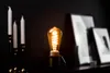 A vintage-style light bulb is illuminated on a dark surface, with candles and framed pictures in the background. Produktbild-Vorschau