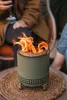 A close-up shot of a Solo Stove Mesa XL Deep Olive fire pit with flames burning, placed on a woven mat. People are visible in the background, suggesting an outdoor social gathering. Náhľad obrázku produktu
