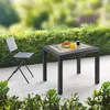 A table and chair set on a patio with a hedge in the background. Produktbild-Vorschau