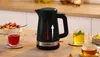 A black electric kettle is shown on a wooden table, surrounded by cups of tea and a bowl of fruit. Anteprima dell’immagine prodotto