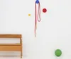 A minimalist scene with a wooden bench, a red jump rope hanging from a blue hook, and a green ball on the floor. Produktbild-Vorschau