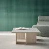 A modern living room setting with a green grid-patterned wall, a plush armchair, and a travertine-look coffee table with an open book. Produktbild-Vorschau