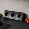 A black power strip with three outlets sits on a black countertop in a kitchen setting, with tomatoes and a bowl of greens nearby. Náhled obrázku produktu