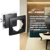 A product shot of a black electrical outlet cover and mounting bracket, with a living room scene in the background. Produktbild-Vorschau