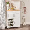 A white kitchen cart with a wooden top, stocked with wine bottles, sits next to a window with curtains. Decorative items like a vase of tulips, a candelabra, and wall art are also visible. Produktbild-Vorschau
