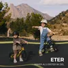 Two children are riding balance bikes on a track with mountains in the background. Produktbild-Vorschau