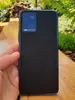 A hand holding a black smartphone with a textured back and a camera module, outdoors with greenery in the background. Produktbild-Vorschau