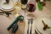 A table setting with wine glasses, a bottle of wine, cutlery, and a Coravin wine system, decorated with pine branches and pinecones for a festive occasion. Podgląd zdjęcia produktu