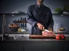 A chef in a denim-like uniform is slicing a piece of meat on a wooden cutting board in a modern kitchen. Produktbild-Vorschau