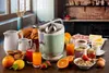 A breakfast spread with a vintage citrus juicer, croissants, cookies, fruit, jam, cereal, and drinks on a wooden table. Produktbild-Vorschau