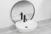 A white oval countertop sink with a black faucet is shown in front of a round mirror, reflecting the sink and faucet. The sink sits on a light gray wooden surface against a white wall. Náhľad obrázku produktu