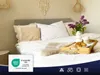 A neatly made bed with white bedding, pillows, and a decorative throw. A wooden tray with a croissant and coffee cup sits on the bed, along with a vase of lilacs. Produktbild-Vorschau