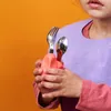 A child wearing a bib and holding a pink utensil holder with a fork and spoon. Voir les images du produit