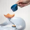 A hand pouring water from a blue cup into a bath toy that has an orange flower-like top and a gray stem with colorful rings at the bottom. Produktbild-Vorschau
