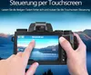 A person is holding a digital camera and interacting with its touchscreen display, which shows a menu with various settings. The background is a scenic landscape with mountains and a lake. Produktbild-Vorschau