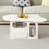 A white coffee table with a unique, wavy shape is placed in a living room setting, in front of a sofa. The table has open shelves and a drawer-like compartment. It is decorated with a bowl of oranges and a small golden bird figurine. Produktbild-Vorschau