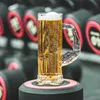 A beer mug with a muscular arm-shaped handle sits on a stand, with blurred dumbbells in the background. Produktbild-Vorschau