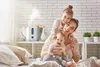 A mother and her two children are sitting on a bed, with a humidifier in the background. Produktbild-Vorschau