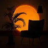 A dimly lit room with a sunset lamp projecting a large orange circle onto a brick wall. A potted plant and an armchair are silhouetted in the foreground. Produktbild-Vorschau