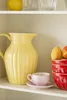 A kitchen shelf with a yellow pitcher, a pink teacup and saucer, and a stack of red bowls with fruit on top. Produktbild-Vorschau