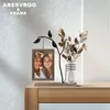 A decorative photo frame with a metal tree branch holding a photo and a glass vase with dried flowers, placed on a wooden table against a textured wall. Produktbild-Vorschau