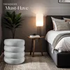 A bedroom scene with a bed, a side table with a lamp, a plant, and a stool. Náhled obrázku produktu