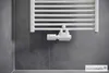 A white smart thermostat is attached to a white radiator against a dark gray tiled wall. Produktbild-Vorschau