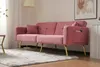 A pink sofa with gold legs is shown in a living room setting with a rug and a lamp. Produktbild-Vorschau