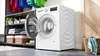 A white washing machine with its door open, in a laundry room setting with shelves of folded laundry and baskets. Anteprima dell’immagine prodotto