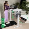 A woman is using a standing desk with a treadmill underneath while on a phone call. The desk is white and has adjustable height features indicated by green arrows. Headphones are hanging from the desk. There are icons on the left side of the image indicati Produktbild-Vorschau