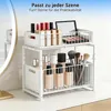 A two-tier white metal organizer shelf holding various makeup products like lipsticks, foundation bottles, and makeup brushes, placed on a white surface next to a mirror. Voir les images du produit