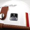 A close-up view of a white motorcycle helmet with a brown leather strap and a sticker indicating size and certification. Podgląd zdjęcia produktu