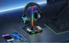 A gaming headset stand with integrated wireless charging for phones, illuminated by RGB lighting, sits on a desk next to a keyboard and monitor. Podgląd zdjęcia produktu